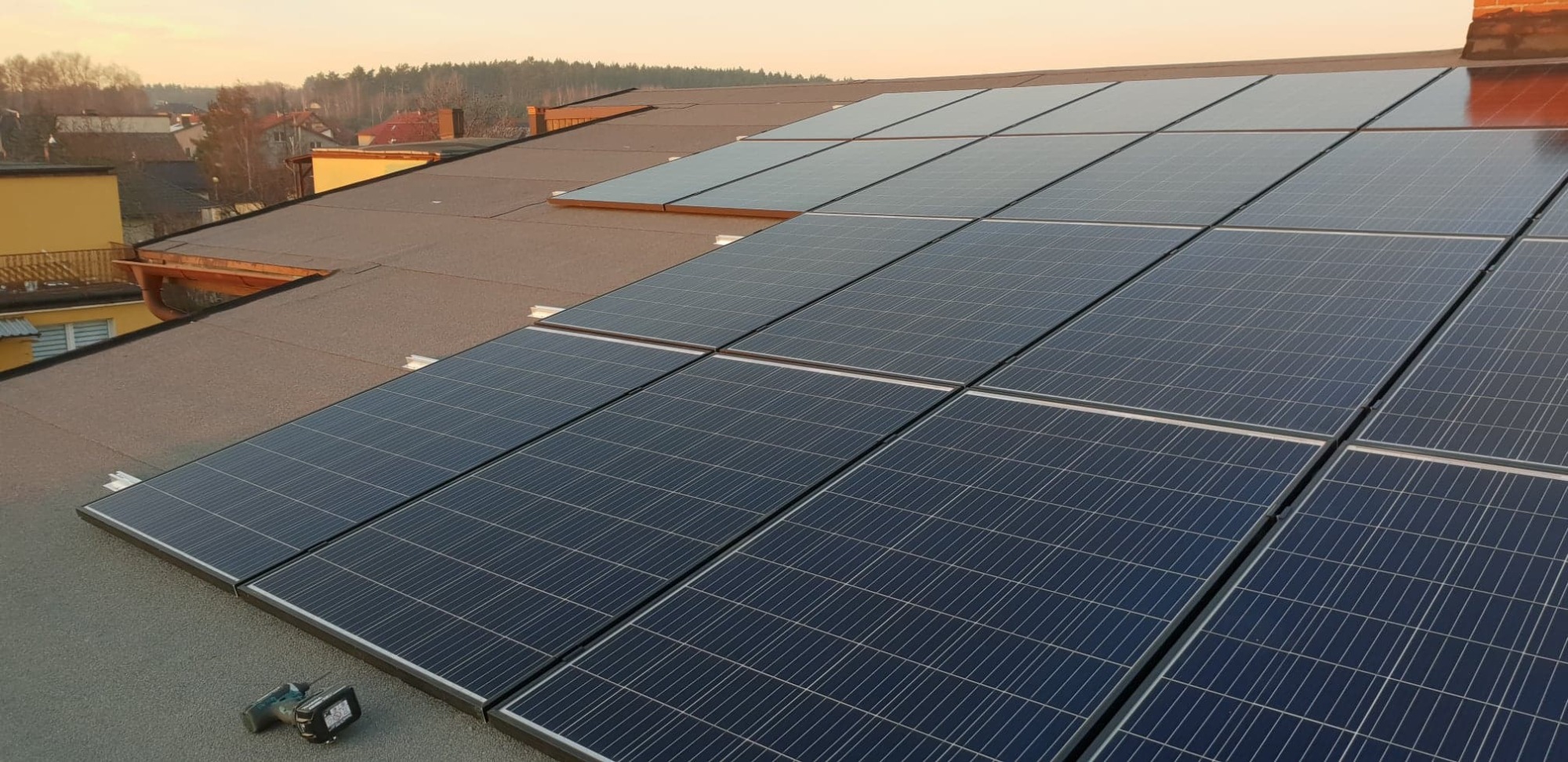 Panele fotowoltaiczne na bitumicznym dachu, z wkrętarką akumulatorową w lewym dolnym rogu i widokiem na zabudowę jednorodzinną w tle.