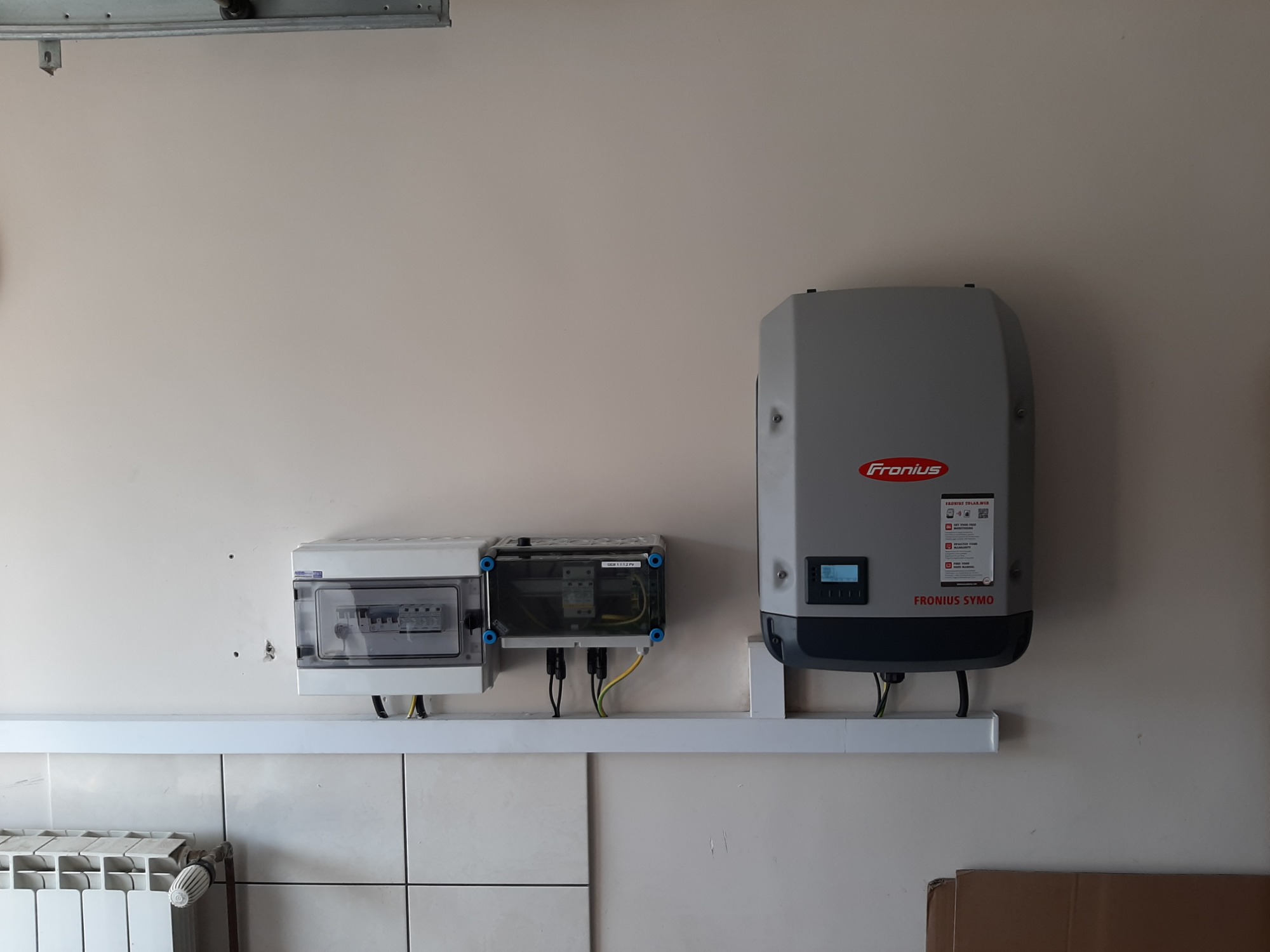 Szare skrzynki elektryczne i inwerter Fronius Symo zamontowane na białej ścianie z kafelkami i grzejnikiem w tle.
