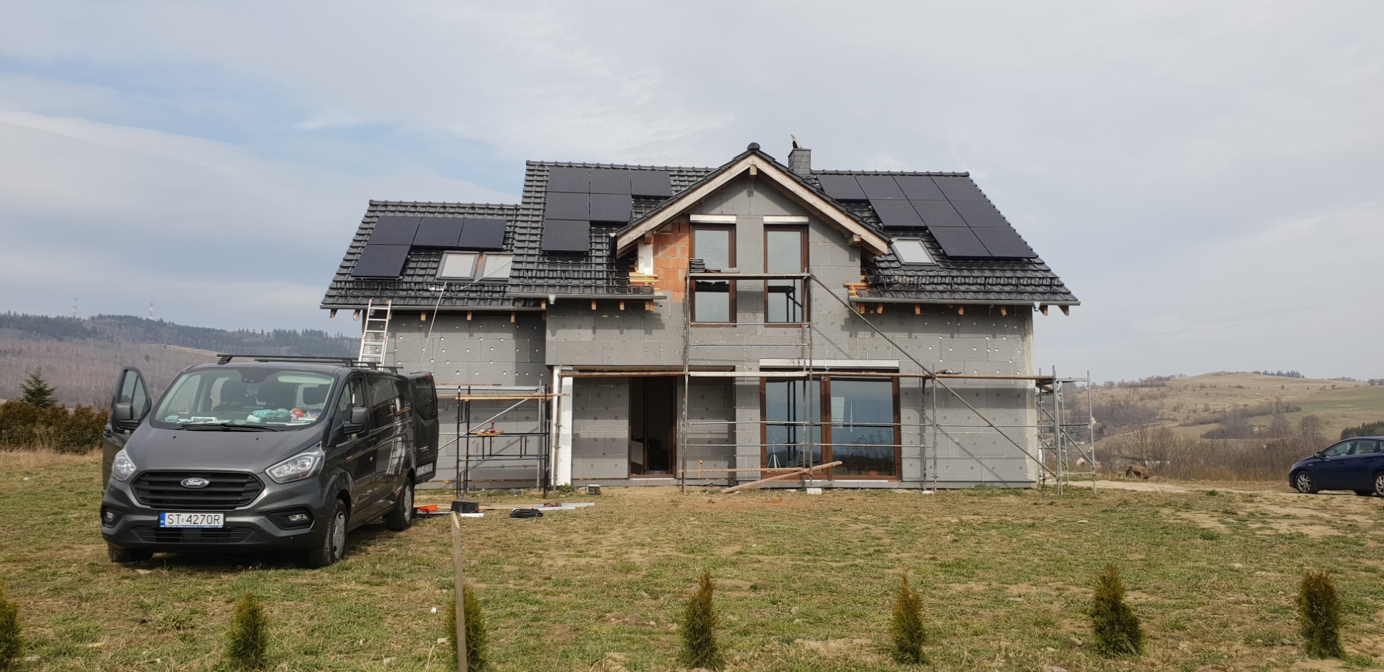 Dom w trakcie budowy z zamontowanymi panelami słonecznymi na dachu, widoczny samochód dostawczy i rusztowania.
