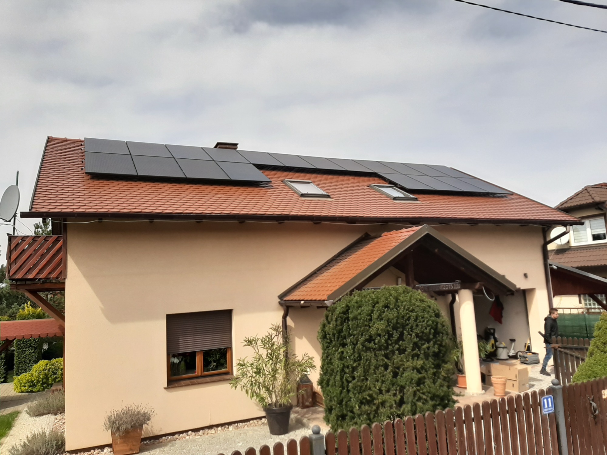 Dom jednorodzinny z zamontowanymi panelami fotowoltaicznymi na dachu z dachówki ceramicznej, widoczne okna dachowe, satelita na balkonie i osoba wchodząca do garażu.