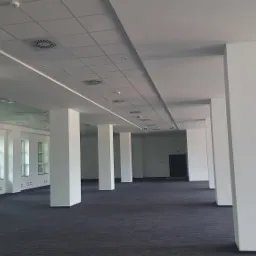 Przestronne, puste biuro z rzędem białych kolumn, podwieszanym sufitem z kratownicą i oświetleniem liniowym LED, widoczne kratki wentylacyjne i czujnik dymu.