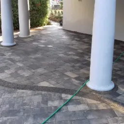 Ułożona kostka brukowa w odcieniach szarości z dekoracyjnym obramowaniem z drobnych kamieni, otaczająca białe kolumny werandy, z widocznym wężem ogrodowym i pojemnikiem z farbą w tle.