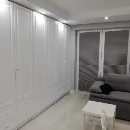 Biała szafa na całej ścianie z frezowanymi frontami i szufladami w dolnej części, widok na salon z szarą sofą i oknem z plisowanymi roletami.