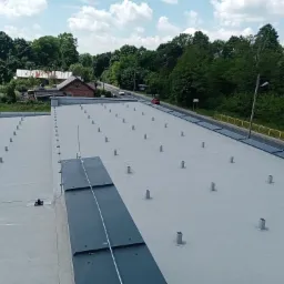 Widok z góry na płaski dach przemysłowy z zamontowanymi elementami instalacji odgromowej, z widocznymi przewodami i uchwytami, w tle drzewa i budynki.