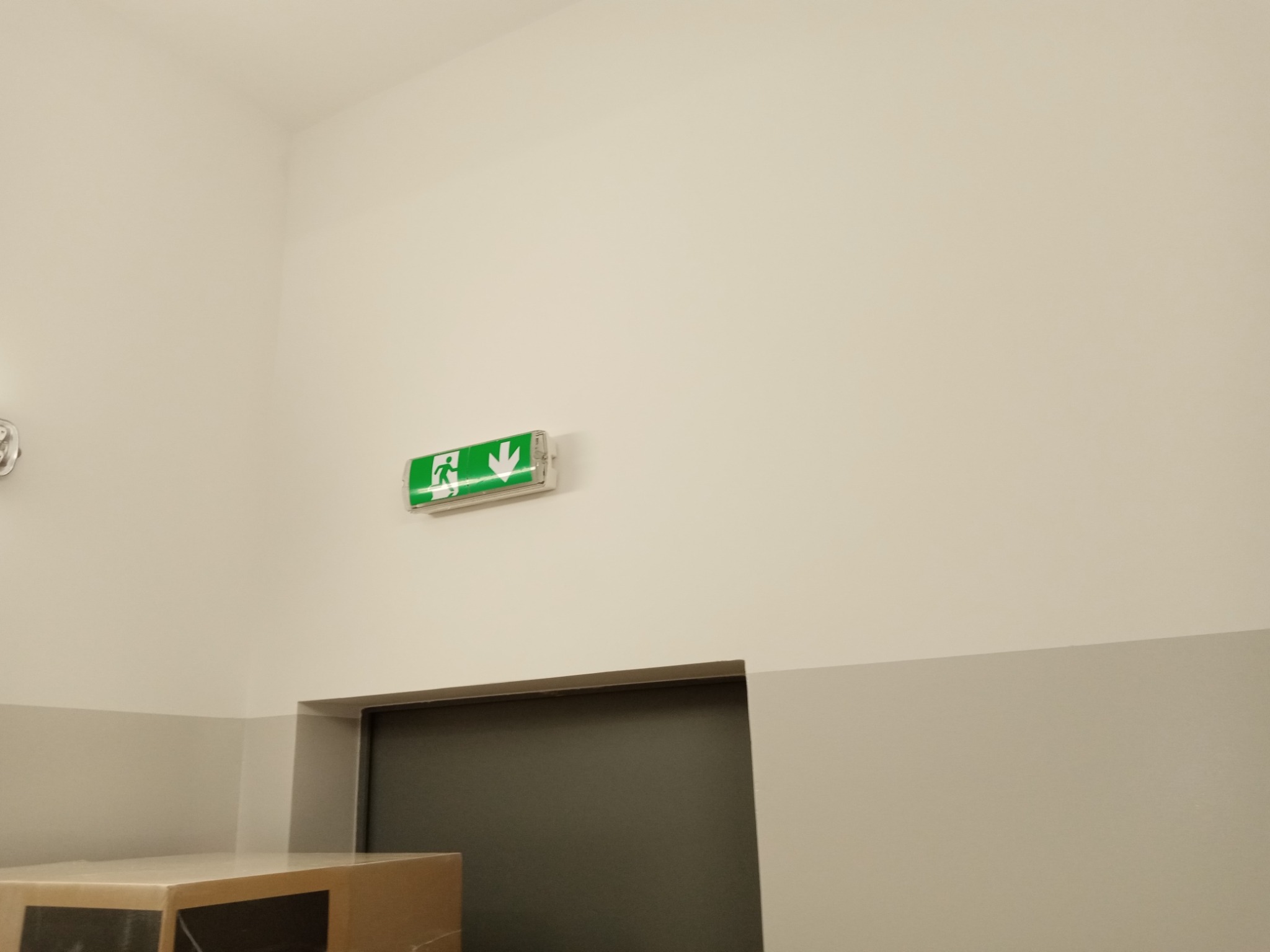 Zamontowany na ścianie, podświetlany znak ewakuacyjny ze strzałką w dół, nad szarymi drzwiami w jasnym pomieszczeniu.