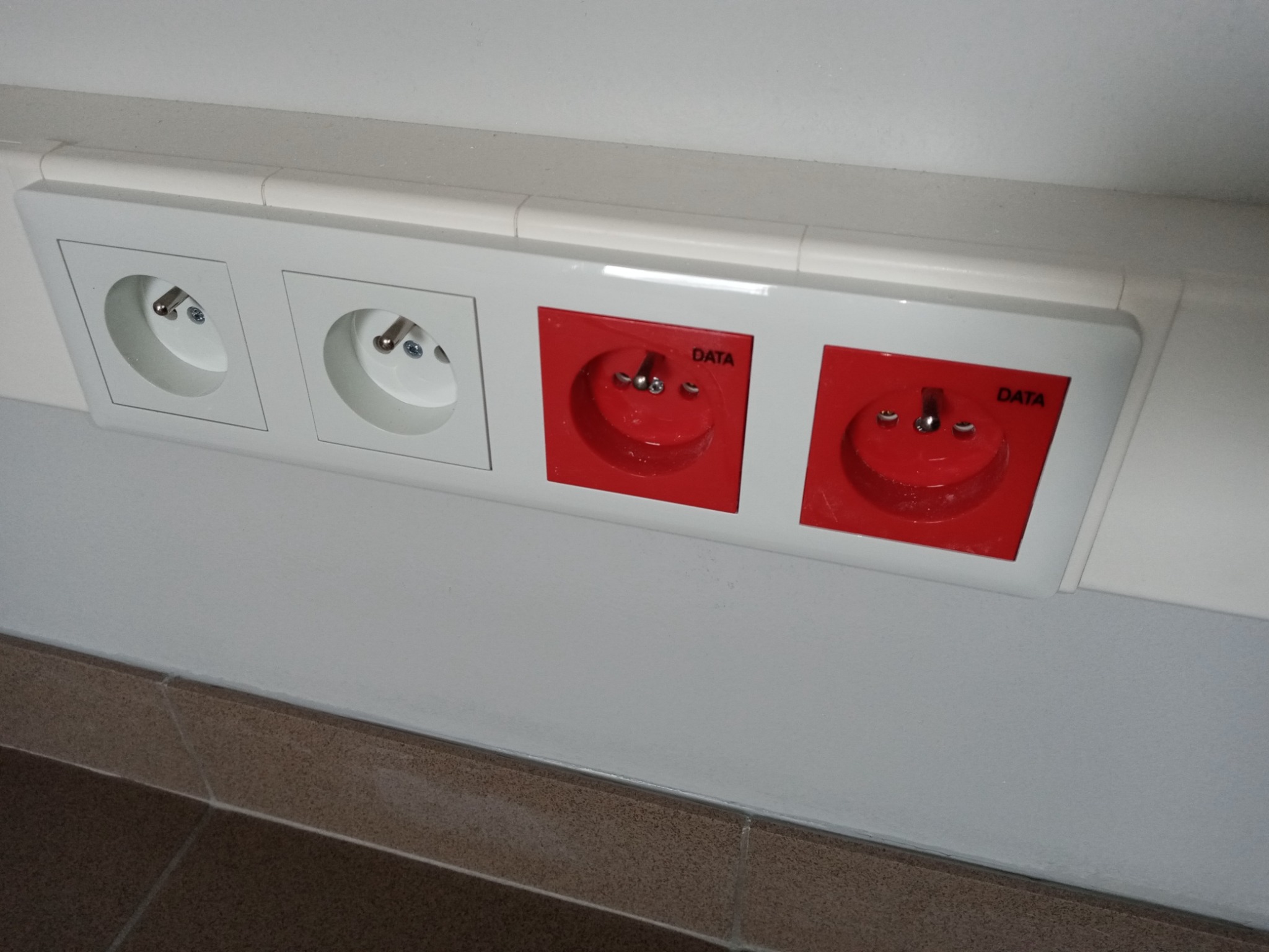 Dwa białe gniazdka elektryczne i dwa czerwone gniazdka DATA zamontowane w białej listwie przypodłogowej na szarej ścianie nad brązowymi płytkami podłogowymi.