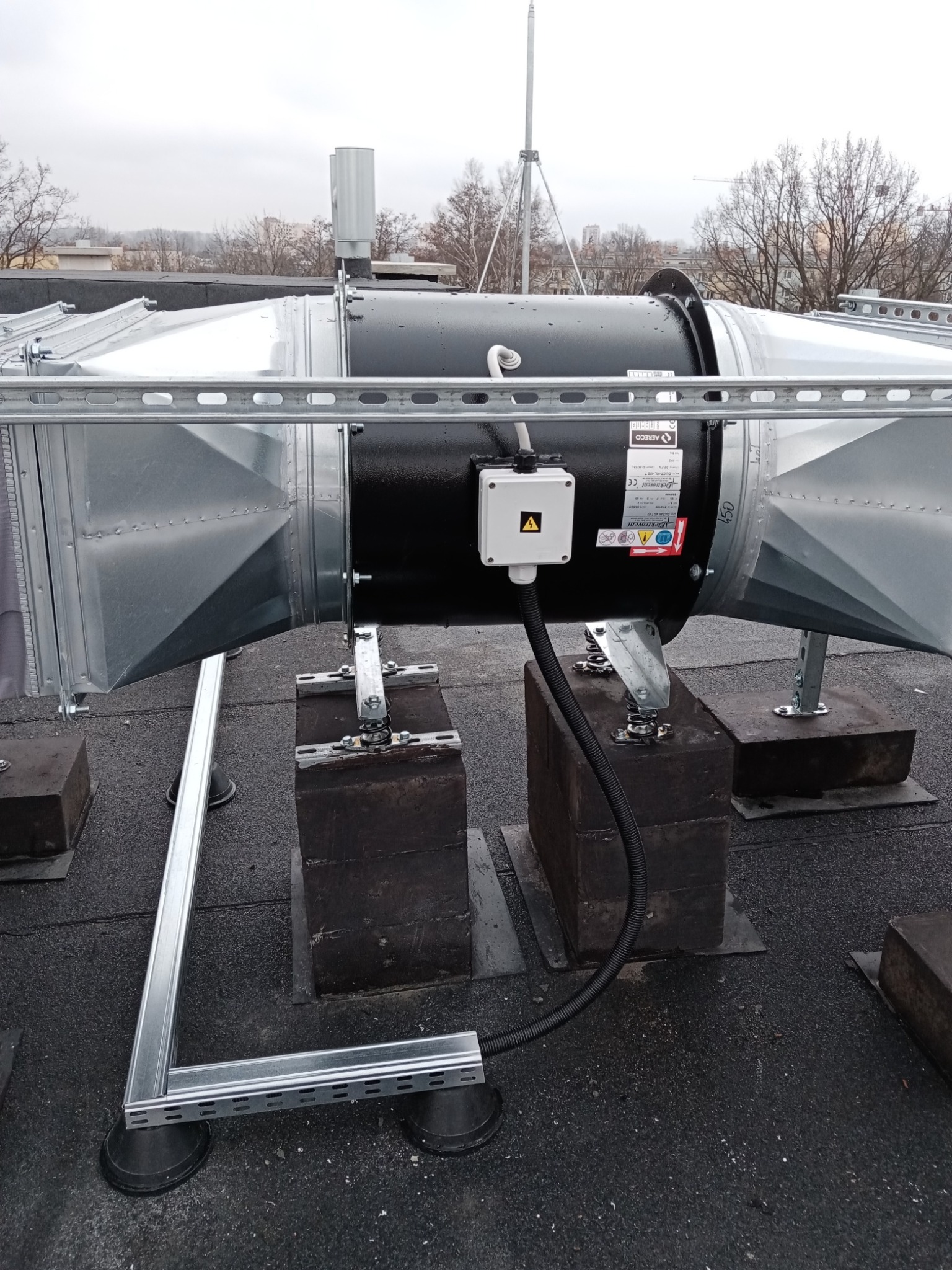 Czarny wentylator przemysłowy z metalowymi kanałami wentylacyjnymi, zamontowany na dachu budynku na betonowych podstawach z amortyzatorami sprężynowymi, widoczna skrzynka elektryczna z symbolem...