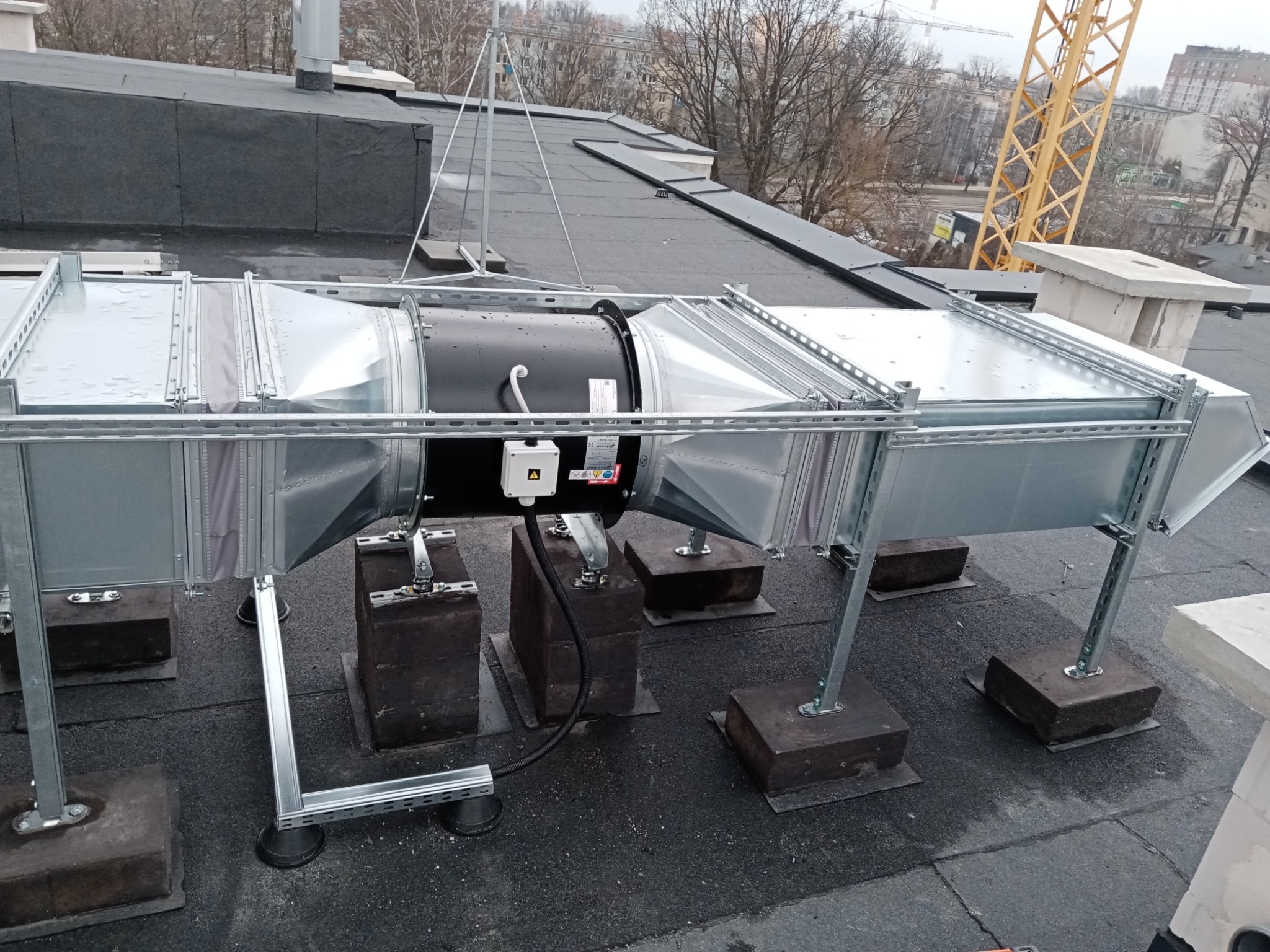 System wentylacyjny z czarnym wentylatorem i srebrnymi kanałami na dachu budynku, zamontowany na betonowych podstawach, z widocznym żurawiem budowlanym w tle.