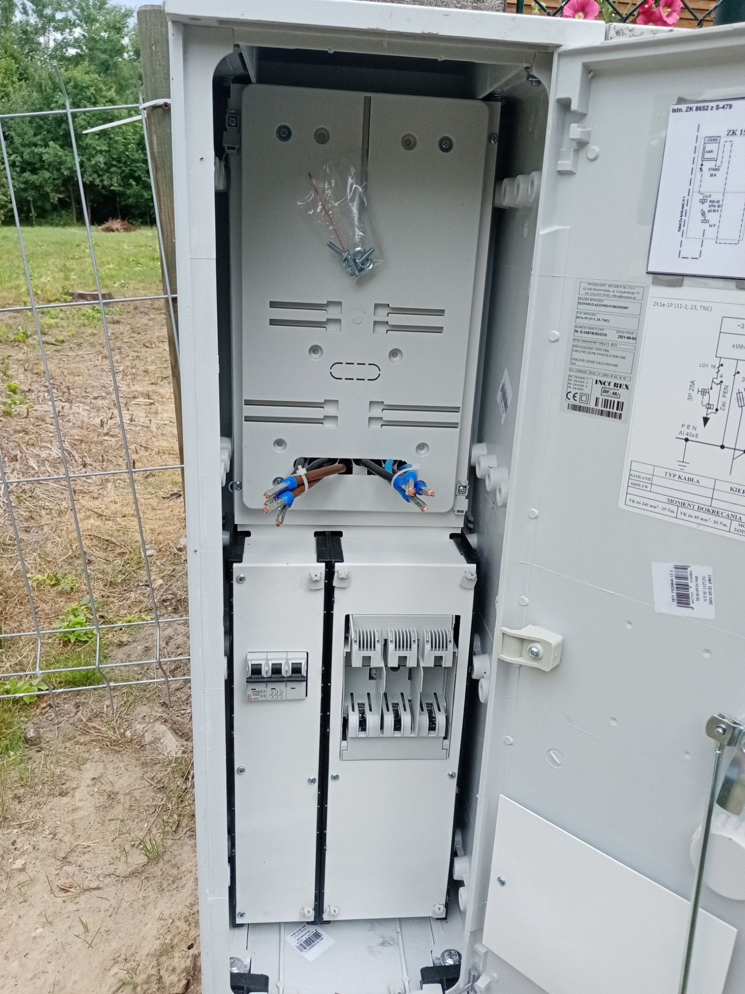 Otwarta skrzynka elektryczna z widocznymi bezpiecznikami, przewodami z końcówkami tulejkowymi i schematem, zamontowana na zewnątrz budynku, obok metalowej siatki ogrodzeniowej.
