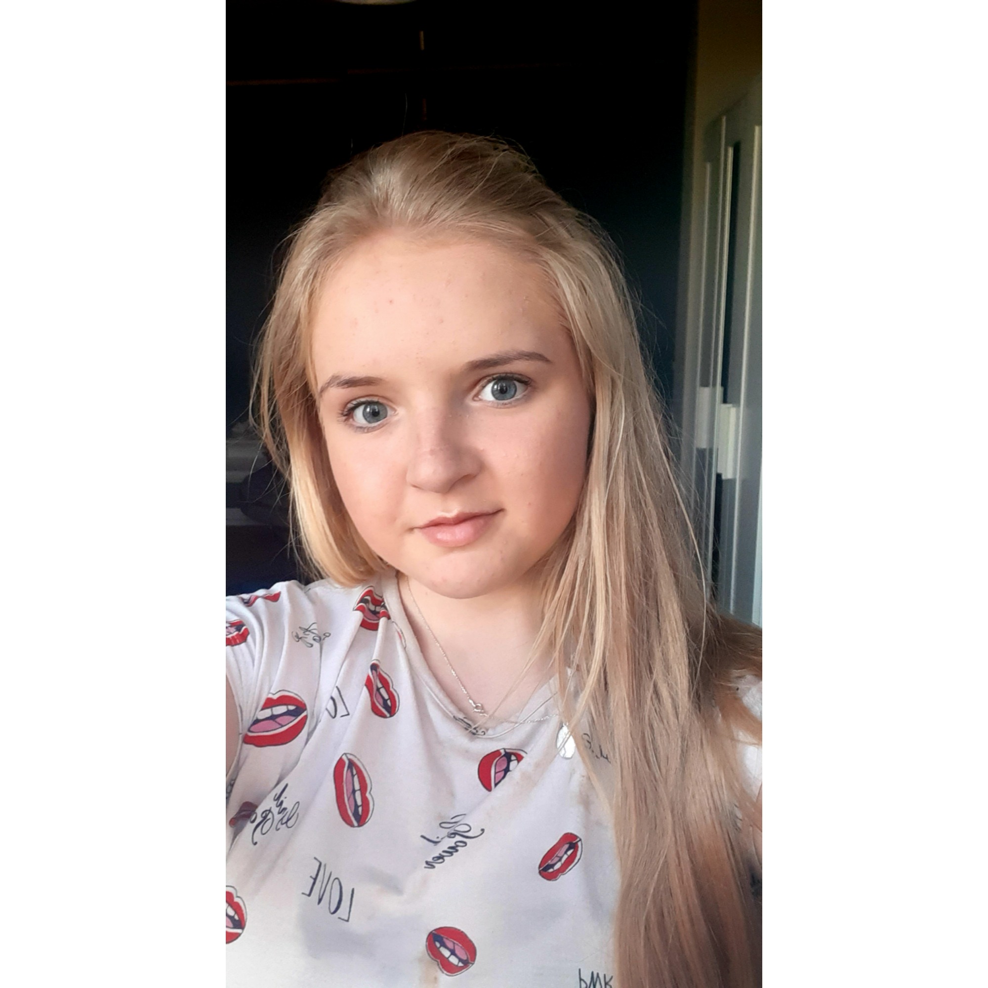 Portret młodej kobiety z blond włosami, ubranej w koszulkę z nadrukiem ust, pozującej w pomieszczeniu z ciemnymi drzwiami w tle.