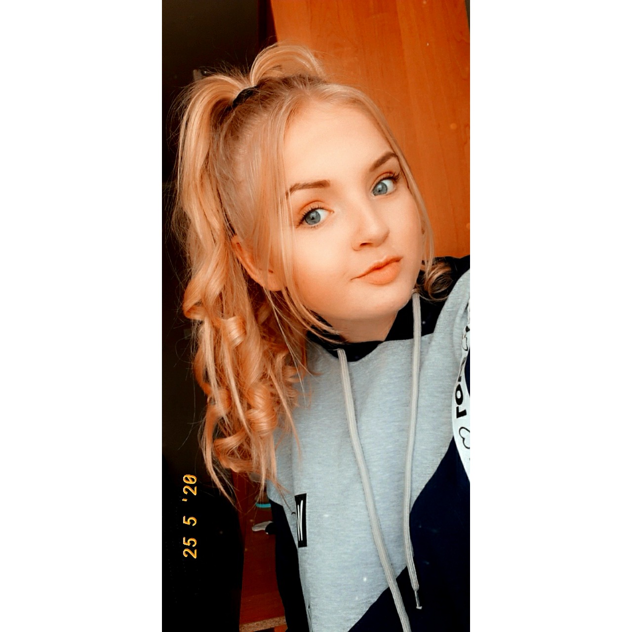 Portret młodej kobiety z blond włosami upiętymi w wysoki kucyk z lokami, ubrana w bluzę z kapturem w kolorach szaro-czarnym, z datą 25.05.20 nałożoną jako filtr.