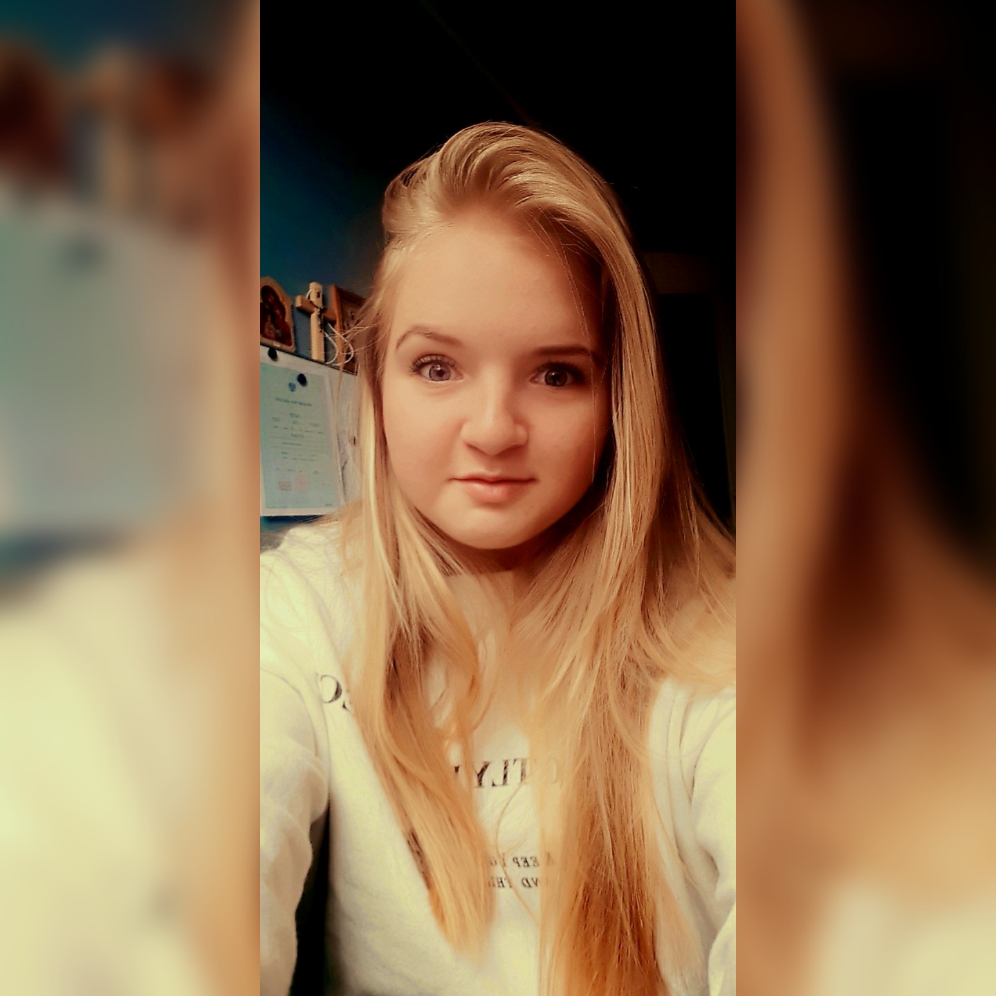 Portret młodej kobiety z długimi blond włosami, ubranej w białą bluzę, z delikatnym uśmiechem, na tle ściany z dekoracjami.