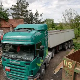 Zielona ciężarówka Scania z białą naczepą typu wywrotka, zaparkowana na placu z widocznym kontenerem na gruz i drzewami w tle.