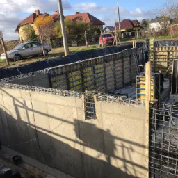 Wykop fundamentowy z częściowo zalanymi betonem ścianami i widocznym zbrojeniem, szalunki w tle, dwa domy jednorodzinne w oddali.