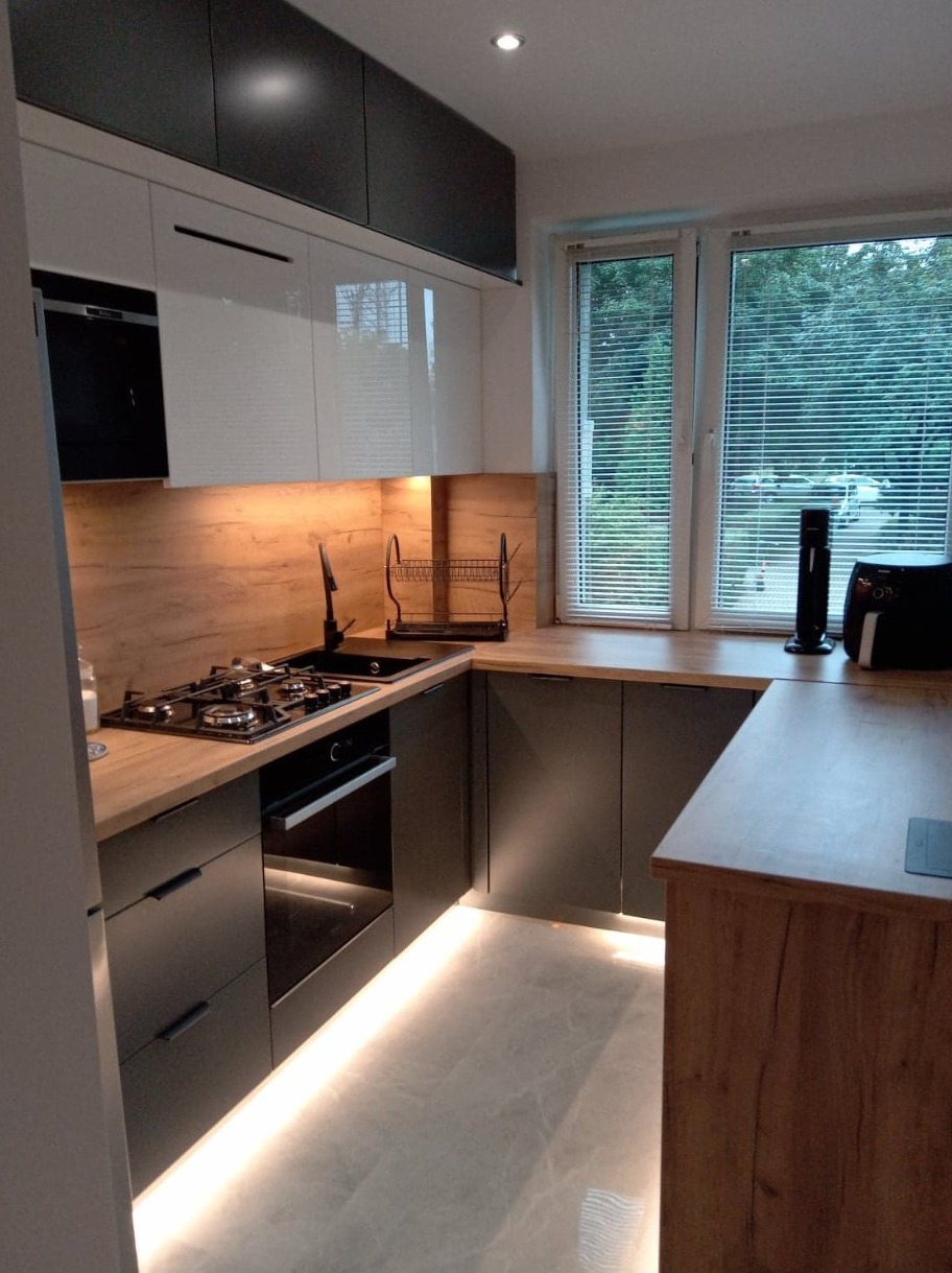 Nowoczesna kuchnia w odcieniach bieli i grafitu z drewnianym blatem, podświetleniem LED pod szafkami i widokiem z okna na zieleń.