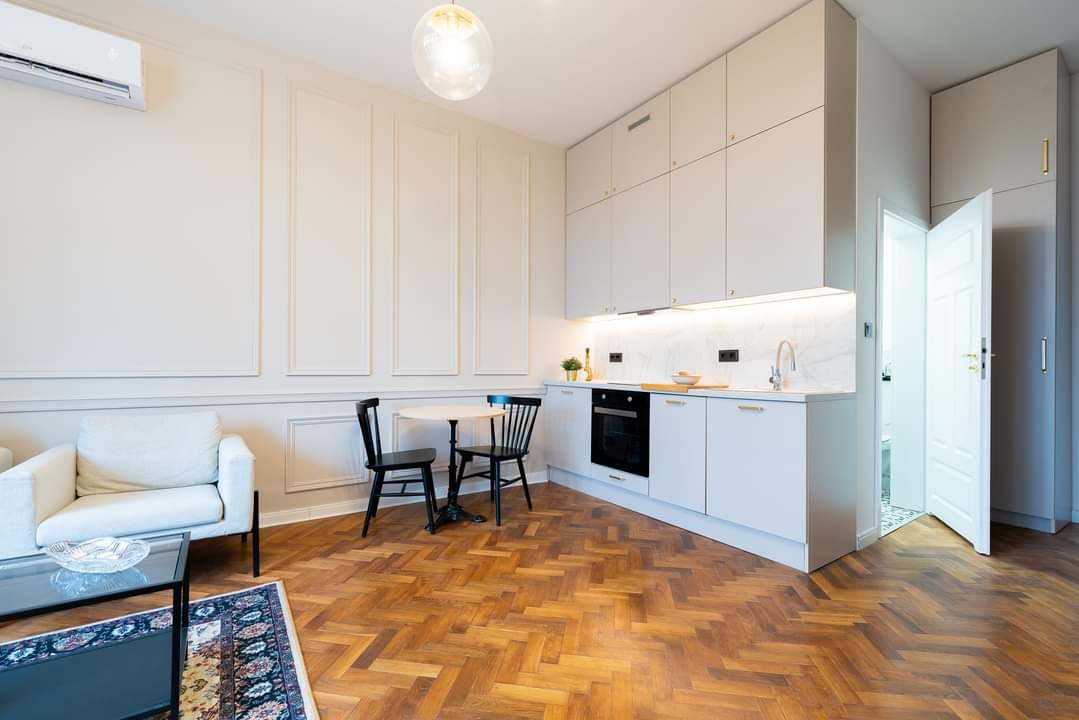 Jasna kuchnia z białymi frontami, złotymi uchwytami i marmurowym blatem, widok na parkiet w jodełkę i salon z kremową sofą.