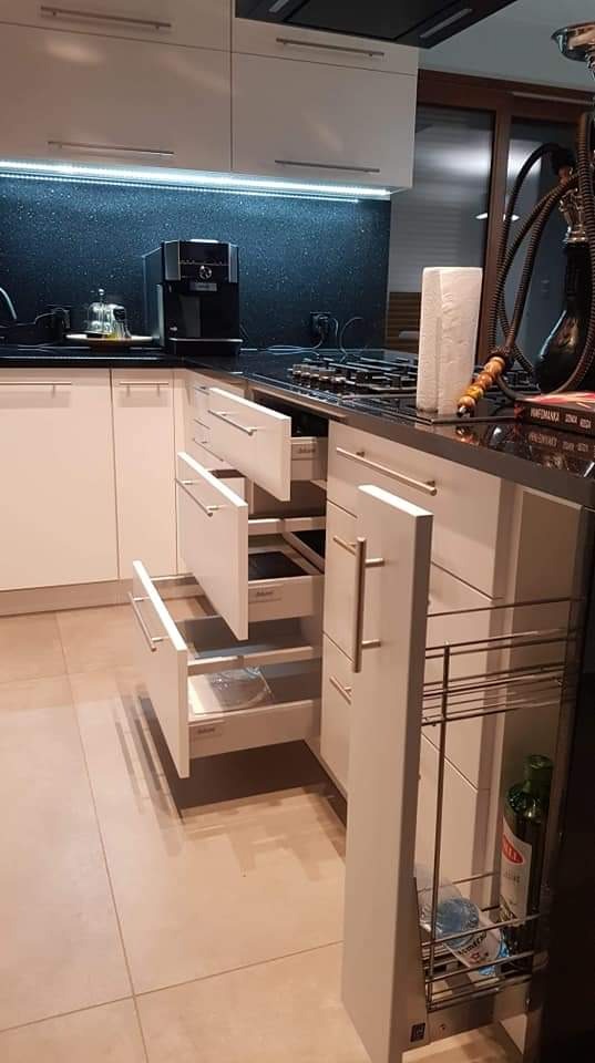 Nowoczesna biała kuchnia z czarnym blatem i błyszczącymi płytkami na ścianie. Wysuwane szuflady z srebrnymi uchwytami, widoczny system cargo z butelką oliwy. Ekspres do kawy na blacie.