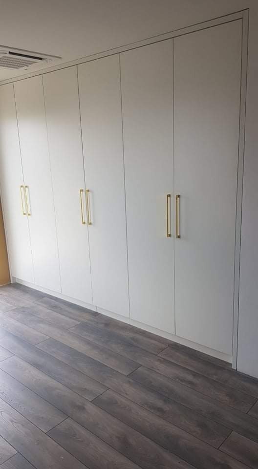 Biała szafa wnękowa z sześcioma drzwiami i złotymi, prostokątnymi uchwytami na tle ciemnej, drewnianej podłogi.