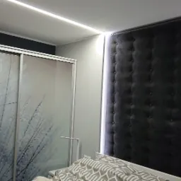 Sypialnia z tapicerowanym, ciemnoszarym panelem ściennym z guzikami, podświetlanym paskiem LED, oraz szafą z przesuwnymi drzwiami z motywem dmuchawca, oświetlona listwą LED.