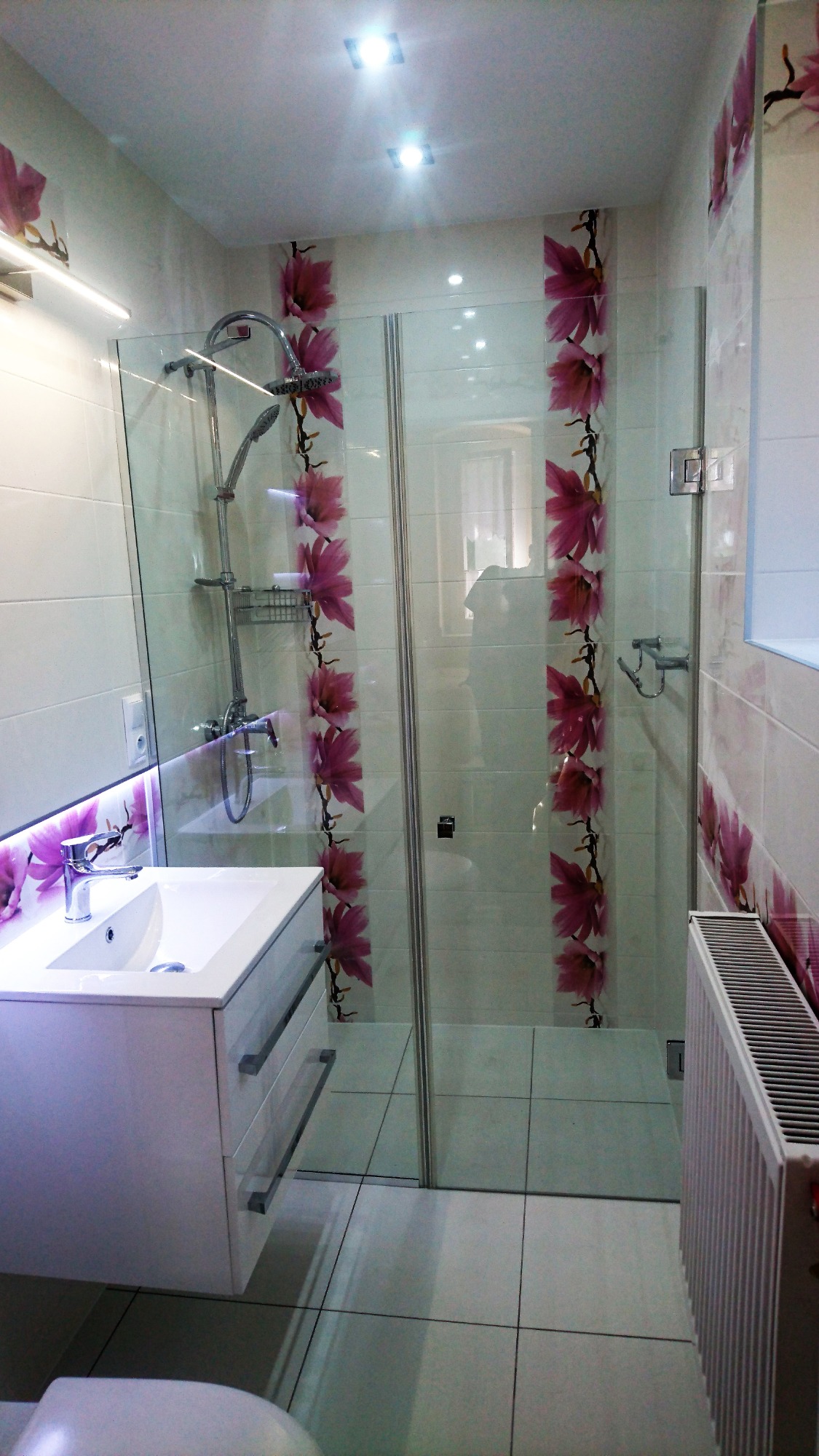 Nowoczesna łazienka z kabiną prysznicową oddzieloną szklaną ścianką z motywem kwiatowym, białą umywalką z szafką i podświetleniem LED, oraz grzejnikiem żeberkowym.