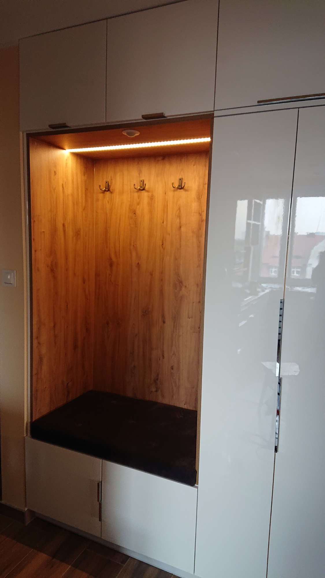 Wnęka w korytarzu z siedziskiem i oświetleniem LED, wbudowana w białą szafę z połyskiem, trzy wieszaki na drewnianym panelu.