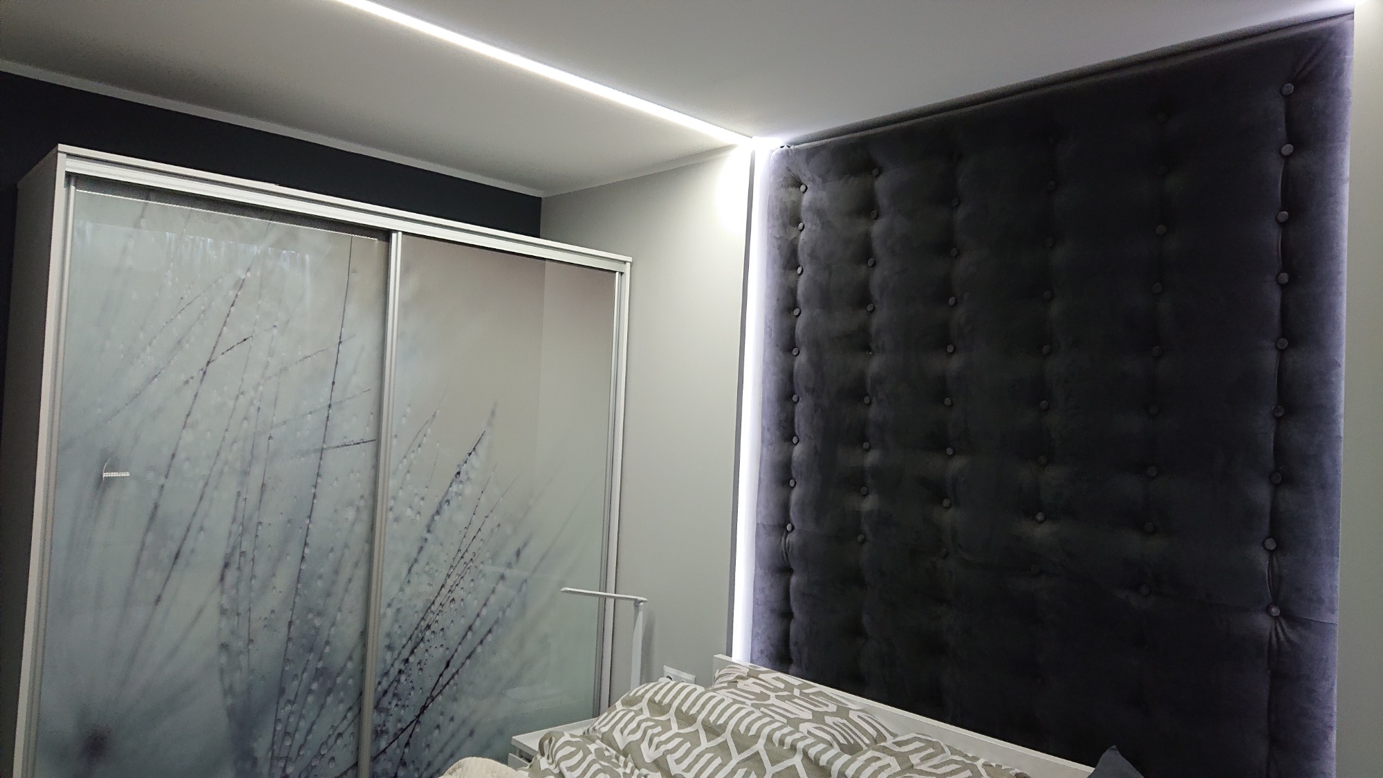 Sypialnia z tapicerowanym, ciemnoszarym panelem ściennym z guzikami, podświetlanym paskiem LED, oraz szafą z przesuwnymi drzwiami z motywem dmuchawca, oświetlona listwą LED.