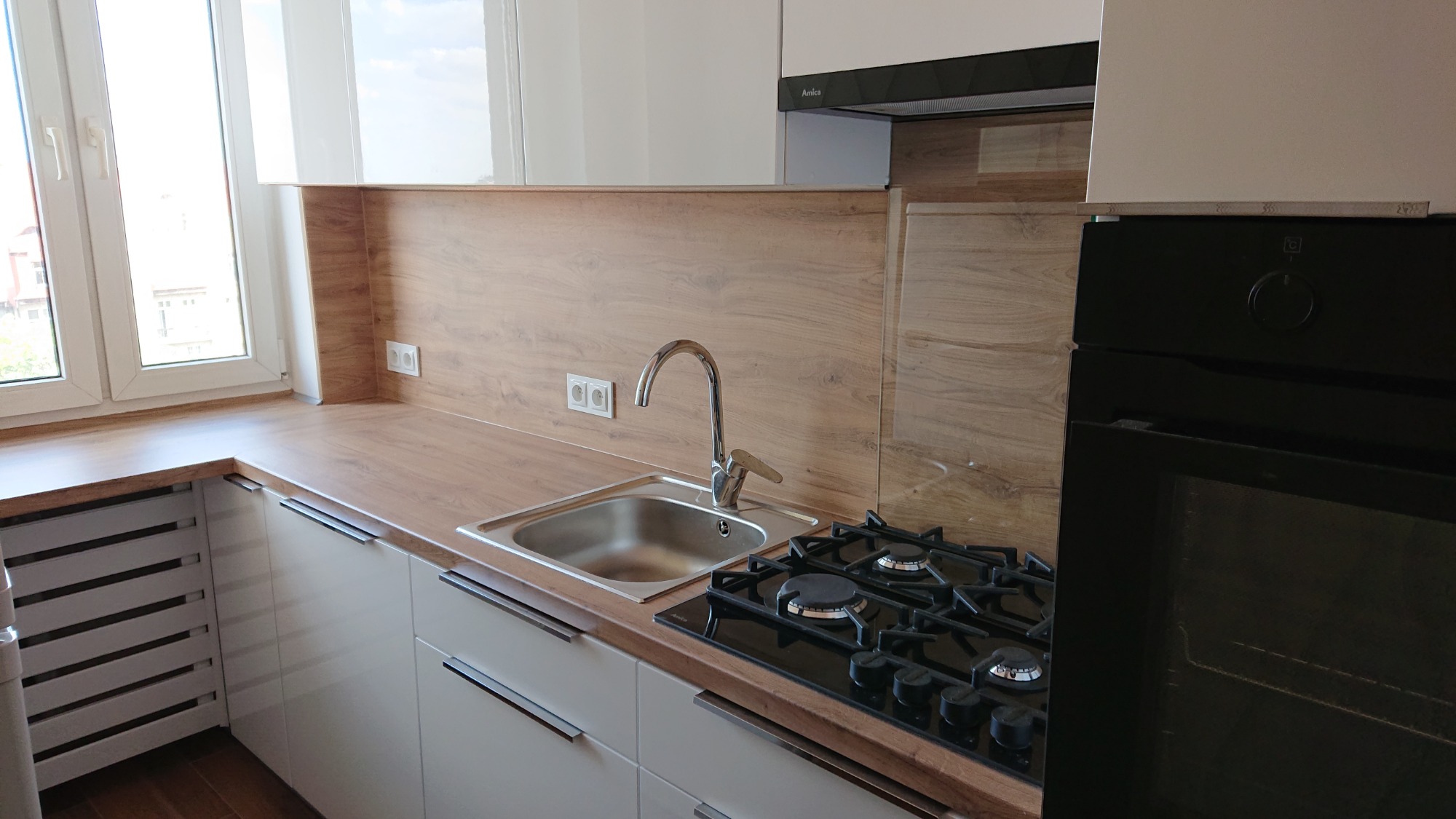 Jasna kuchnia z drewnianym blatem, chromowanym zlewem i czarną płytą gazową oraz piekarnikiem, widok na okno z białymi ramami.