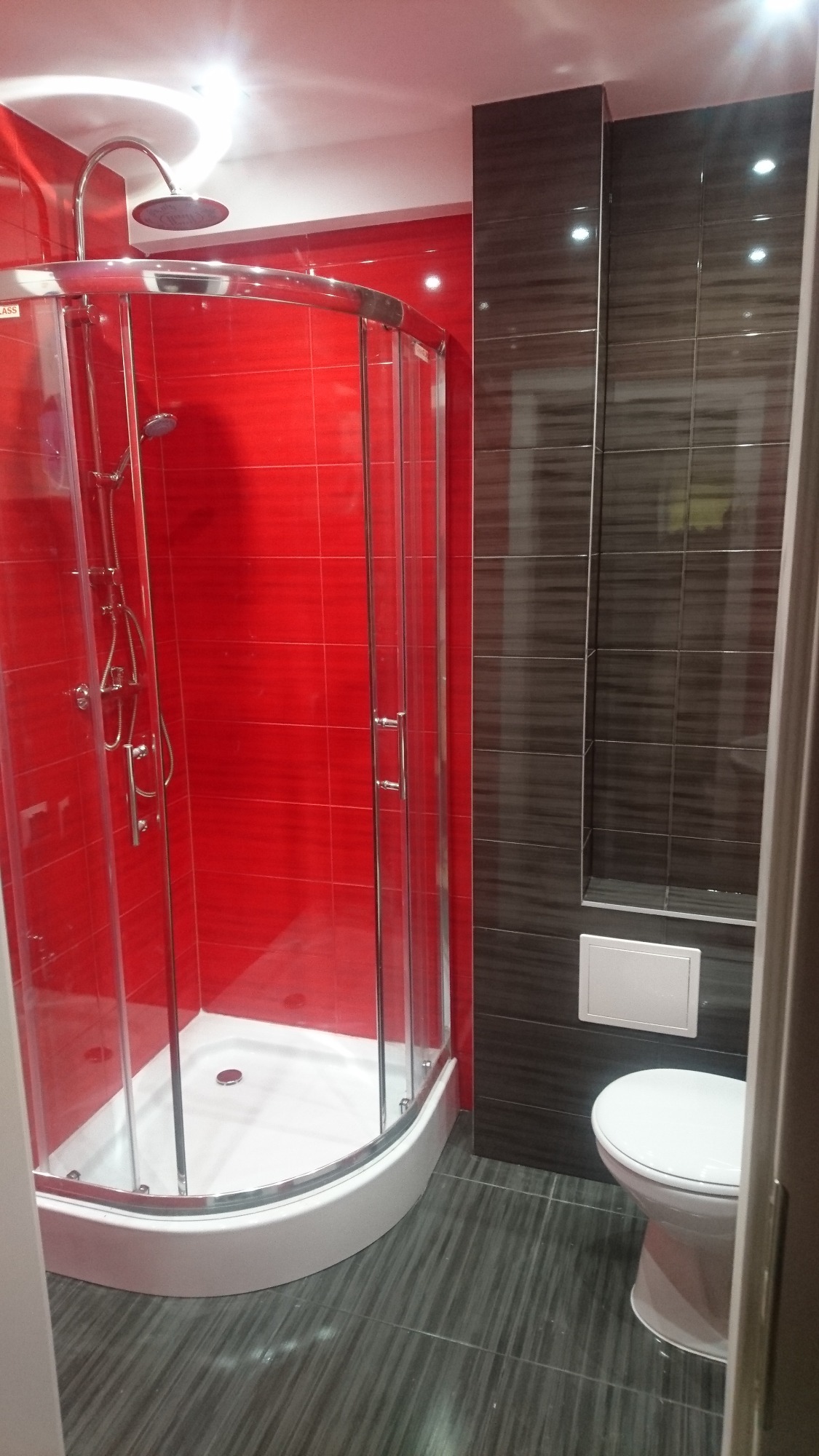 Ukończona łazienka z kabiną prysznicową z chromowanymi elementami na tle czerwonych płytek, toaleta w rogu, szare płytki podłogowe i ciemne płytki na ścianie.