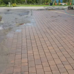 Częściowo wyczyszczona kostka brukowa na podjeździe, widoczna różnica w kolorze między czystą i brudną powierzchnią, na pierwszym planie plandeka ochronna, w tle myjka ciśnieniowa i wąż.