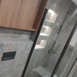 Grupa Handlowo - Usługowa Nalborski - Kajetan Nalborski - Nowoczesna łazienka z szarymi płytkami imitującymi marmur, kabiną prysznicową z czarnymi profilami, wnęką z półkami podświetlonymi od góry oraz drewnianą szafką wiszącą.