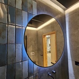 Grupa Handlowo - Usługowa Nalborski - Kajetan Nalborski - Łazienka z błyszczącymi, niebiesko-szarymi płytkami, okrągłym lustrem z podświetleniem LED i widocznym fragmentem wnętrza przez otwarte drzwi.