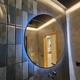 Łazienka z błyszczącymi, niebiesko-szarymi płytkami, okrągłym lustrem z podświetleniem LED i widocznym fragmentem wnętrza przez otwarte drzwi.