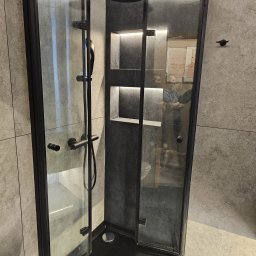 Grupa Handlowo - Usługowa Nalborski - Kajetan Nalborski - Nowoczesna kabina prysznicowa z czarnymi profilami, deszczownicą i wnękami z podświetleniem LED, widoczna w odbiciu sylwetka osoby i fragment toalety.