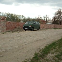 Grupa Handlowo - Usługowa Nalborski - Kajetan Nalborski - Zielony bus firmy budowlanej zaparkowany na brukowanej drodze przed murem z czerwonej cegły, w tle drzewa i pochmurne niebo.