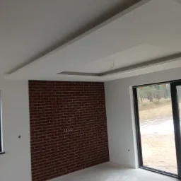 Wnętrze pokoju z fragmentem ściany wyłożonym czerwoną cegłą, białe ściany i sufit z dekoracyjnym podwieszanym elementem, duże okno tarasowe z widokiem na las.