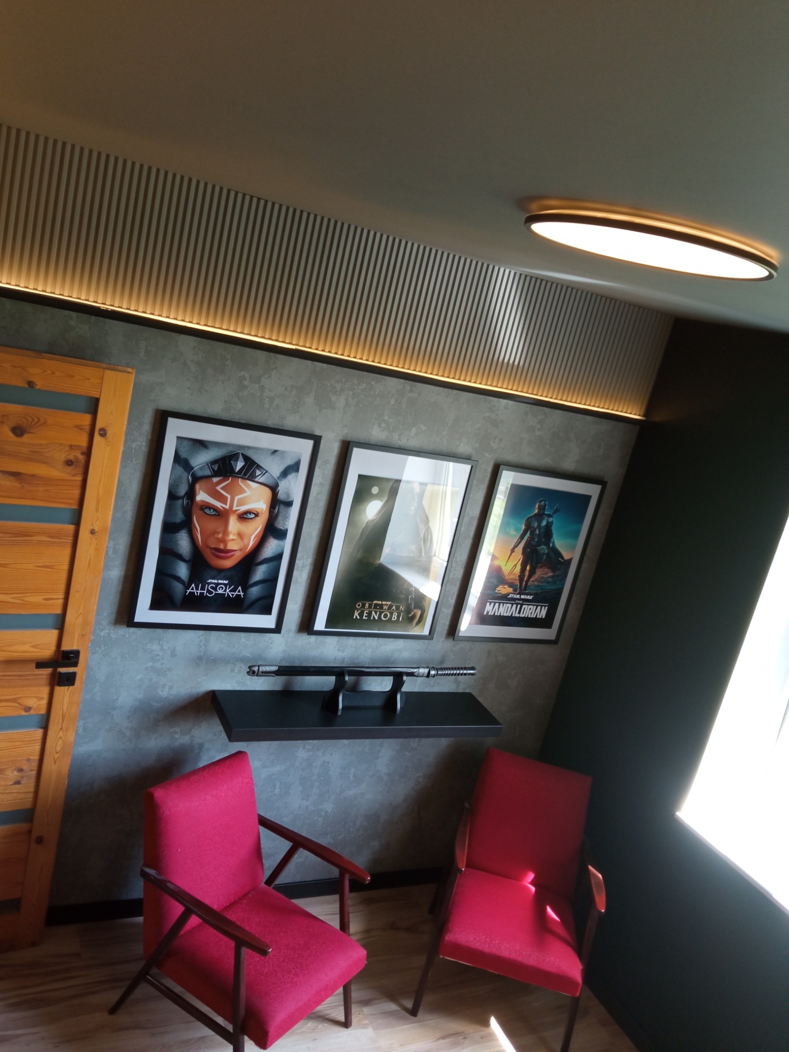 Pokój z drewnianymi panelami na suficie, oświetleniem LED, plakatami z Gwiezdnych Wojen, półką z mieczami świetlnymi i dwoma czerwonymi fotelami.