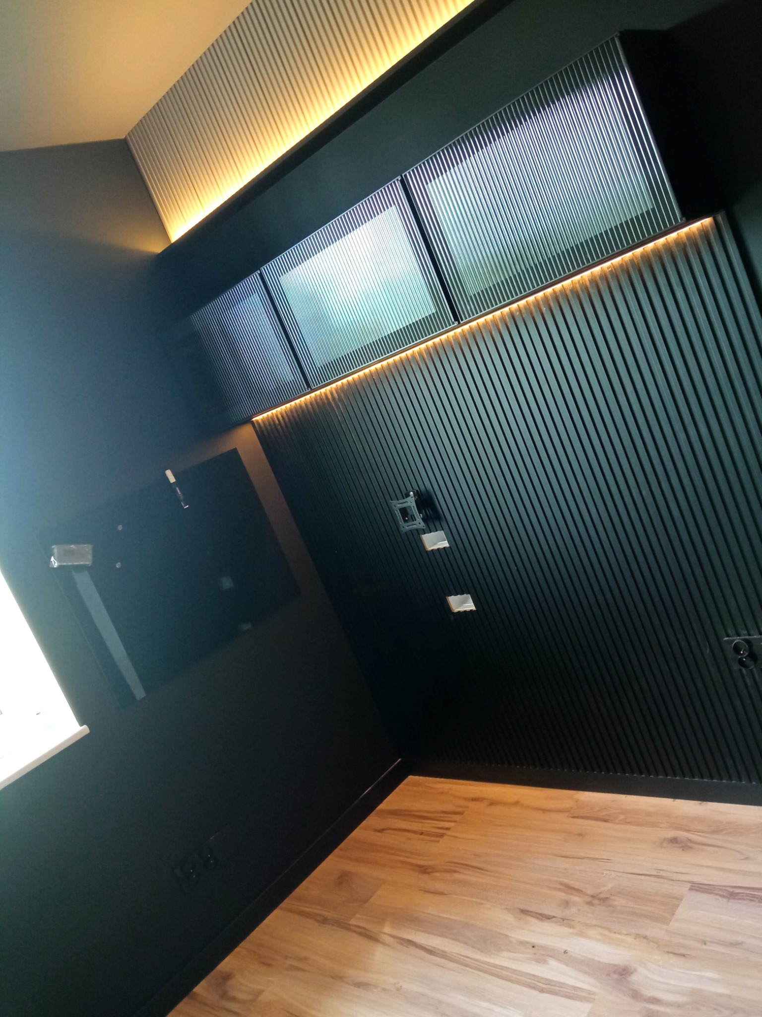 Nowoczesne wnętrze z czarnymi, pionowo żebrowanymi panelami ściennymi i podświetleniem LED, drewnopodobna podłoga, uchwyty do montażu telewizora na ścianie.