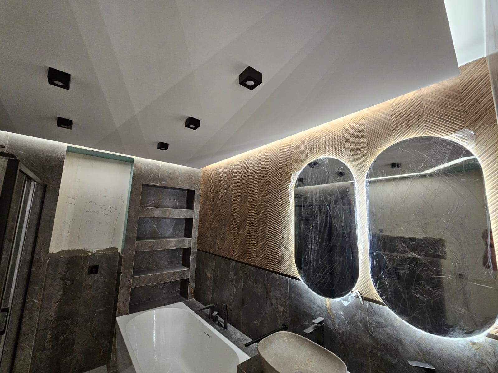 Nowoczesna łazienka w trakcie wykańczania: marmurowe płytki, podświetlane lustra owalne w folii ochronnej, wnęki półkowe, czarne oświetlenie sufitowe.