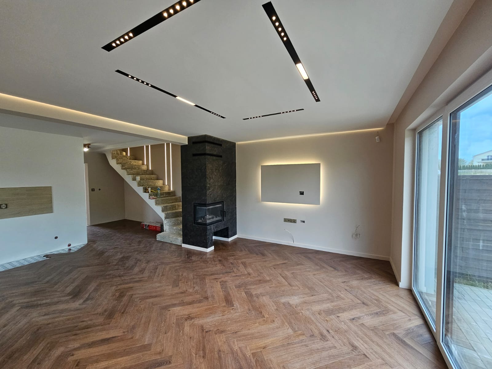 Salon po remoncie z parkietem ułożonym w jodełkę, czarnym kominkiem obłożonym kamieniem i nowoczesnym oświetleniem sufitowym.