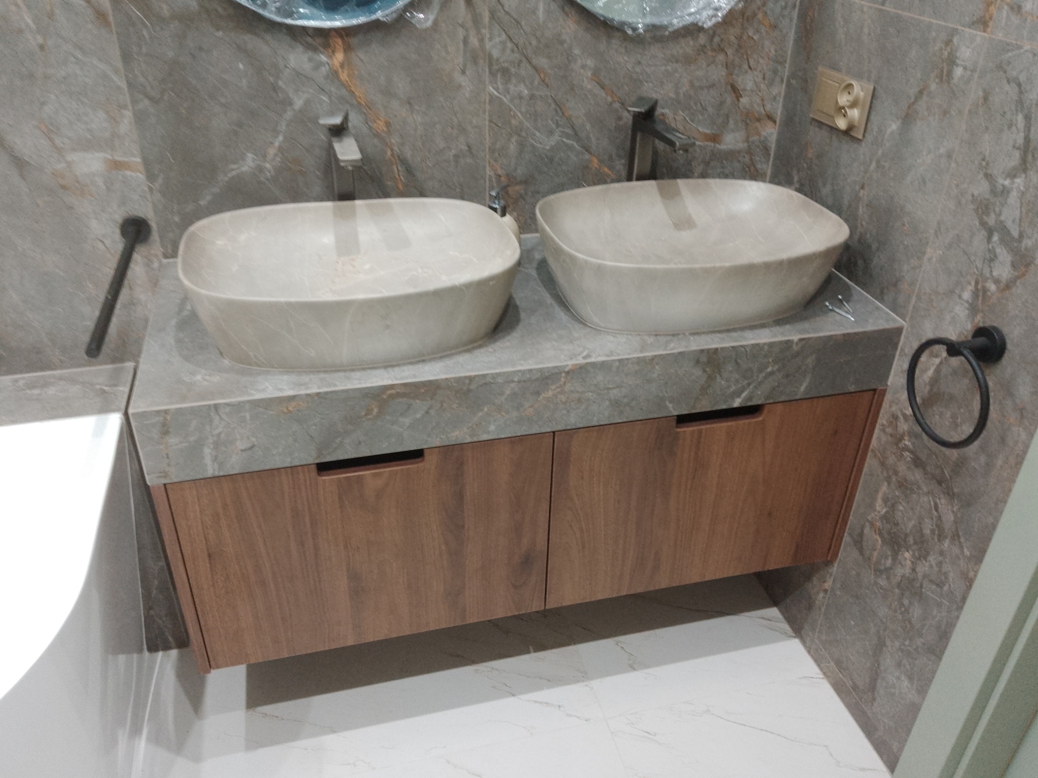Dwie beżowe umywalki nablatowe o owalnym kształcie, z nowoczesnymi, minimalistycznymi kranami, osadzone na blacie z szarego marmuru z brązowymi żyłkami, szafka pod umywalką z drewnianymi frontami...