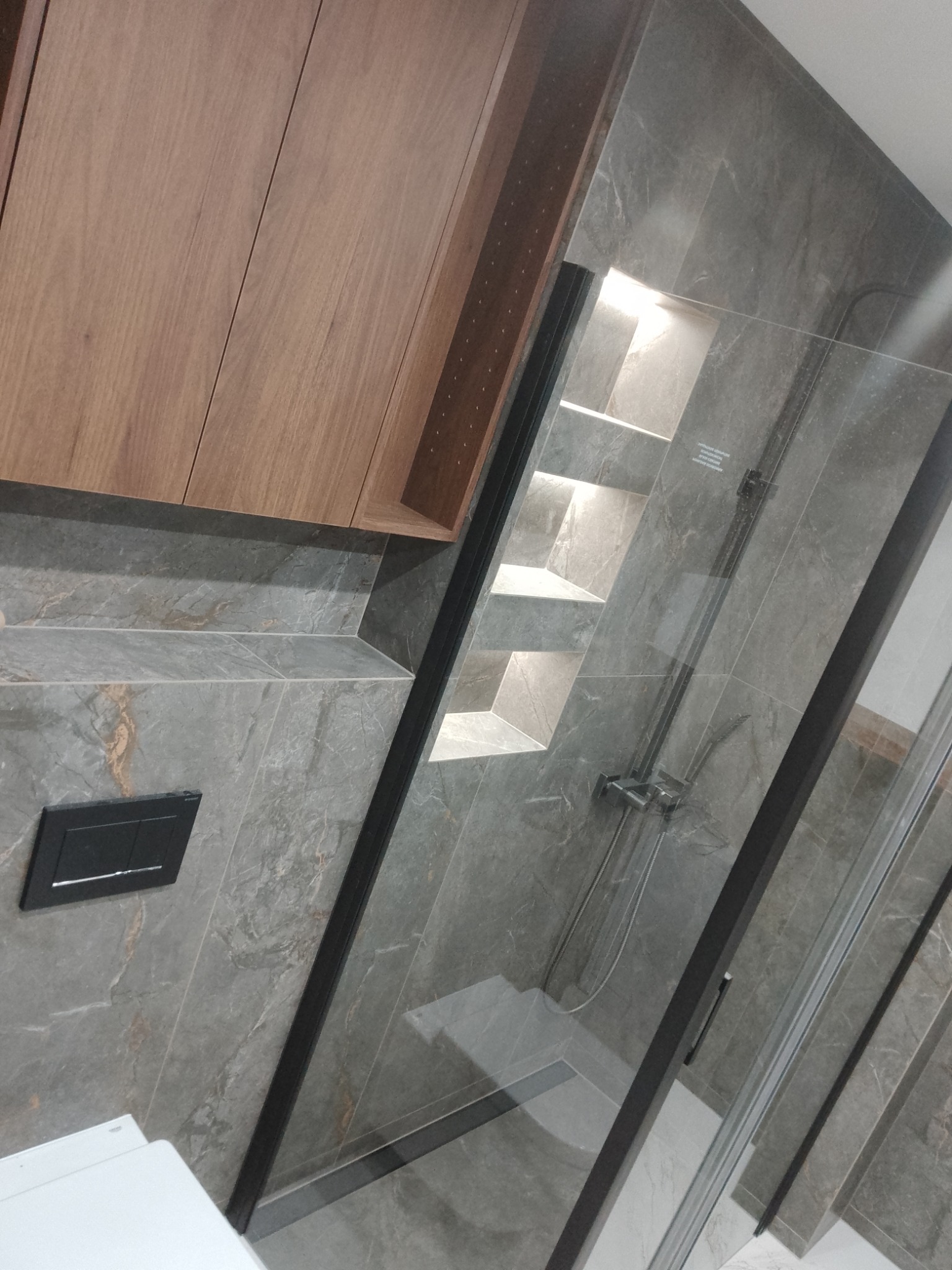 Nowoczesna łazienka z szarymi płytkami imitującymi marmur, kabiną prysznicową z czarnymi profilami, wnęką z półkami podświetlonymi od góry oraz drewnianą szafką wiszącą.