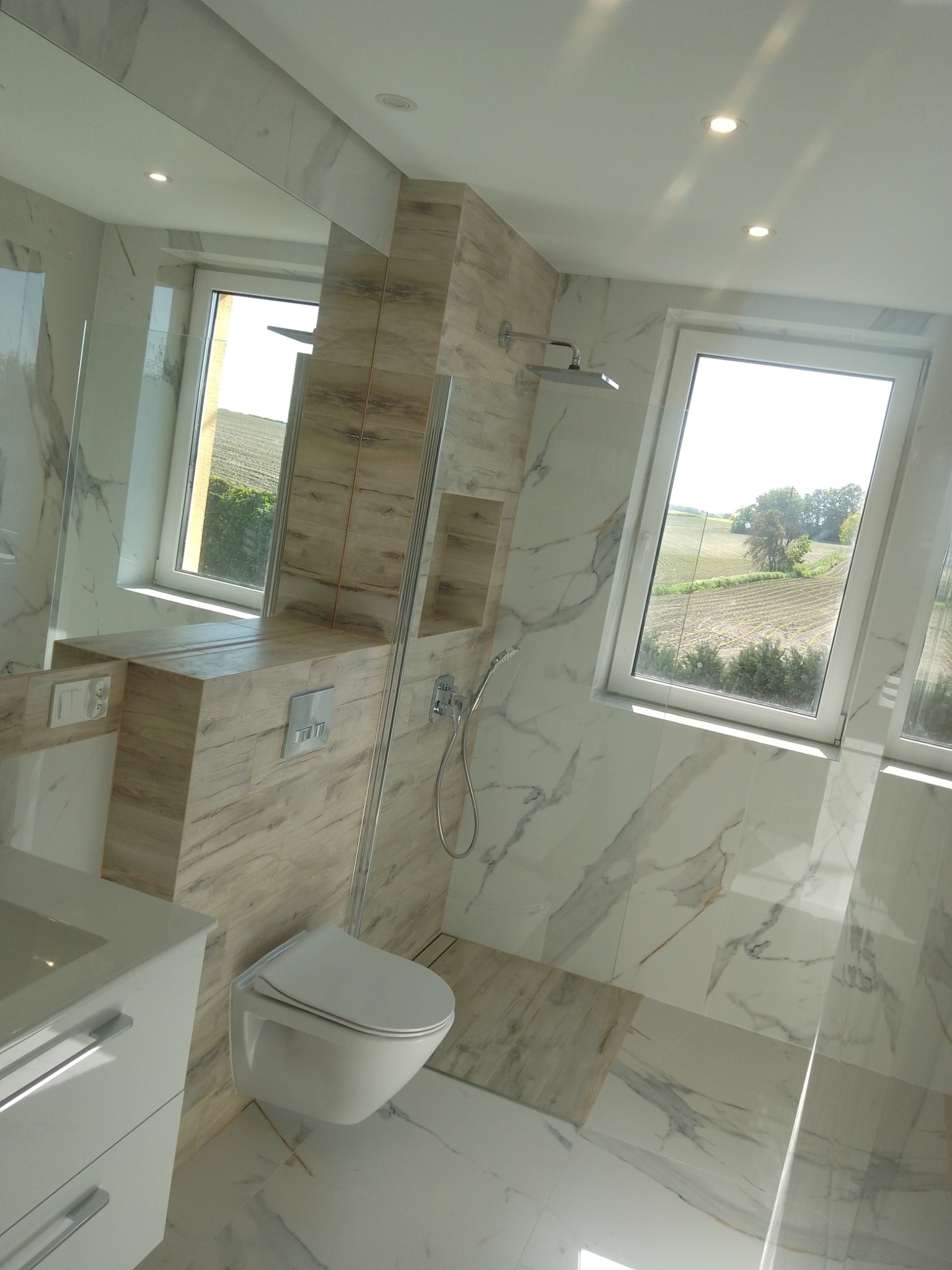 Nowoczesna łazienka z białymi marmurowymi płytkami, podwieszaną toaletą i widokiem na pole z okna.