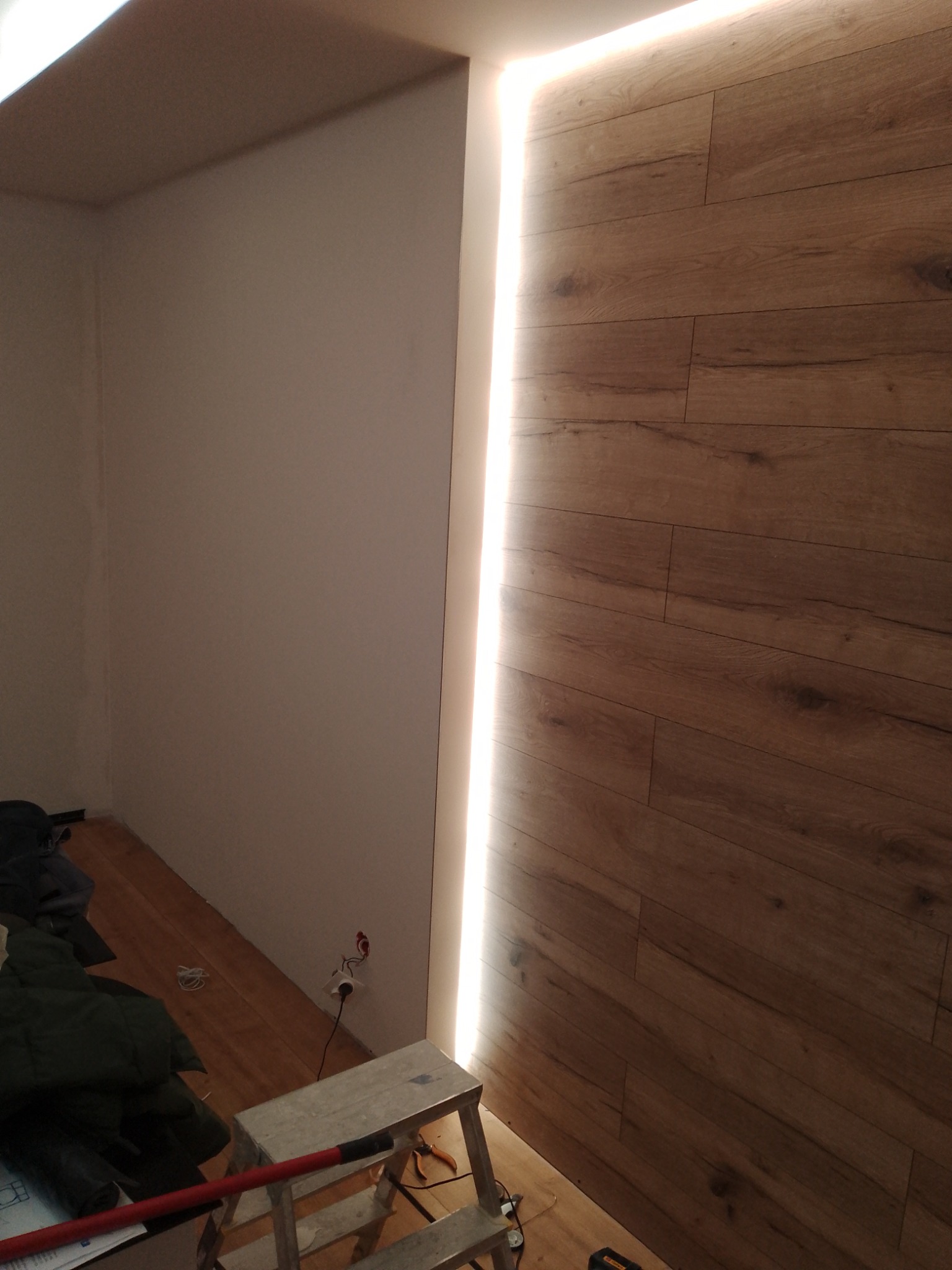 Pokój w trakcie prac: Częściowo wykończona ściana z paneli drewnopodobnych z oświetleniem LED, surowe ściany, drabina malarska i narzędzia na podłodze.