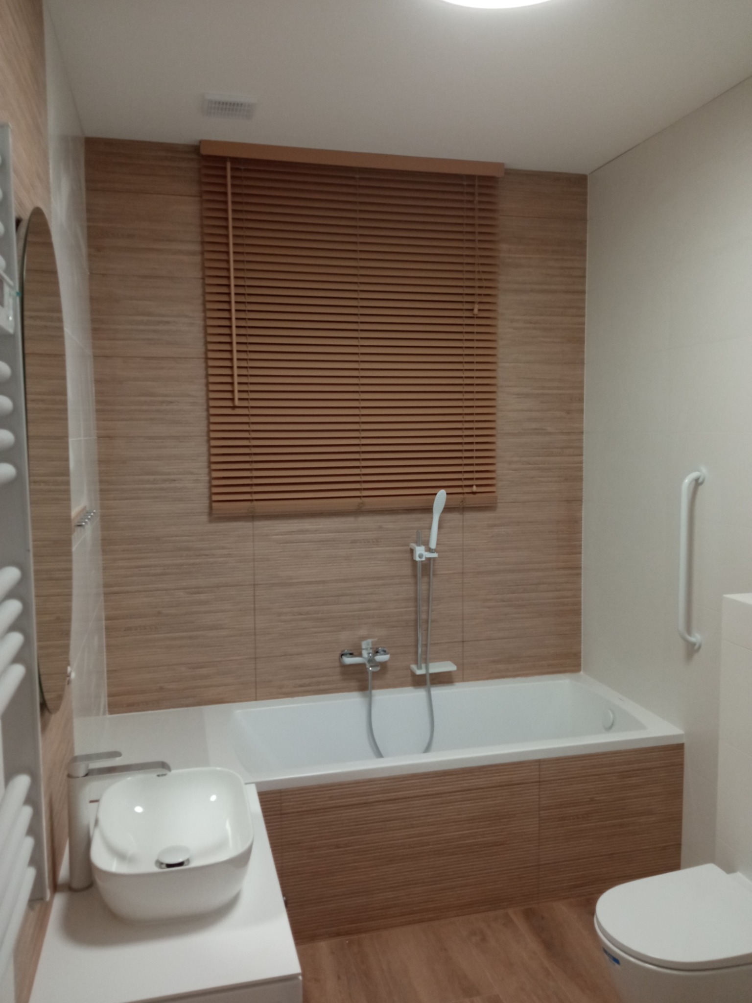 Jasna łazienka z drewnopodobnymi płytkami, białą wanną, umywalką nablatową i drewnianymi żaluzjami w oknie.