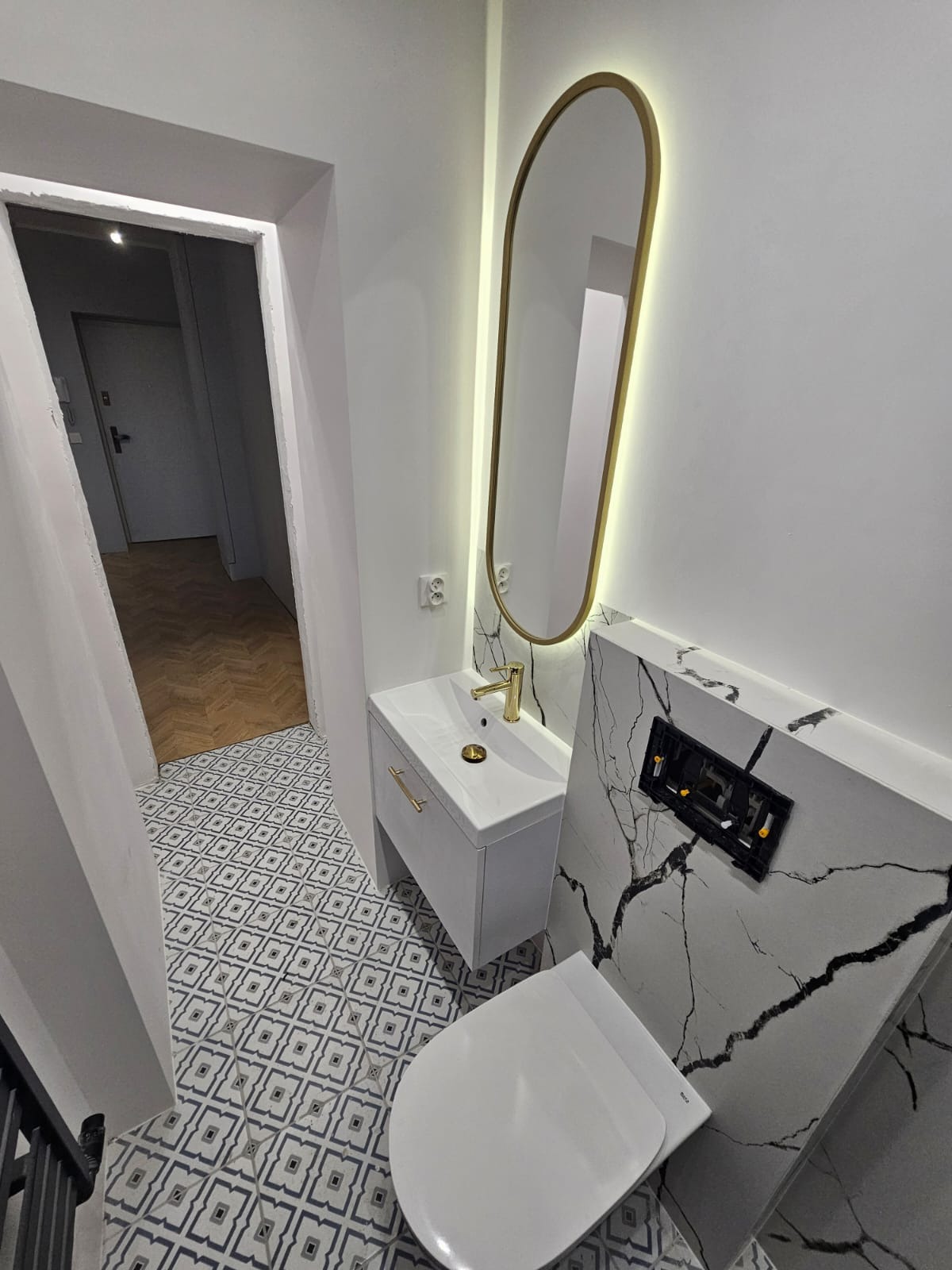 Wykończona łazienka z podwieszaną toaletą, umywalką z szafką, złotą armaturą i podświetlanym lustrem owalnym; na podłodze wzorzyste płytki, w tle widoczny parkiet w jodełkę.