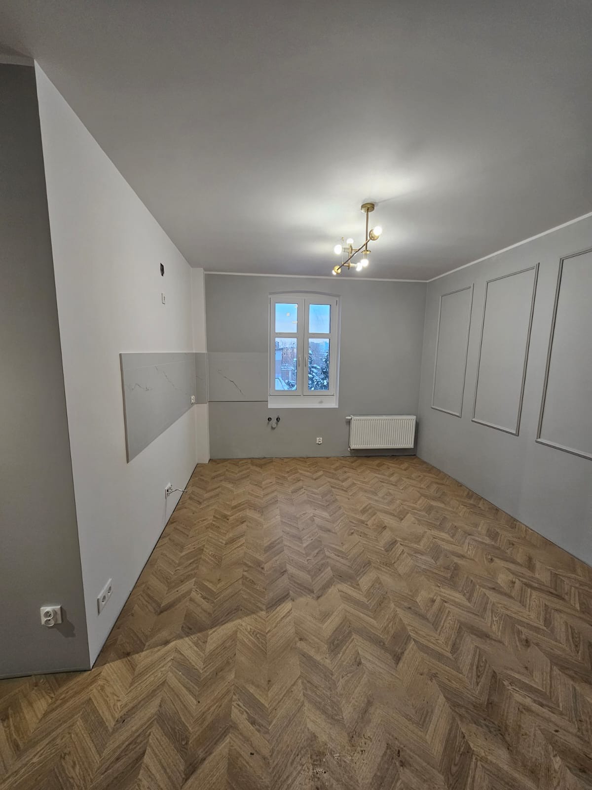 Pusty pokój po remoncie z parkietem ułożonym w jodełkę, szarą ścianą z dekoracyjnymi ramkami, białym grzejnikiem i fragmentem ściany z płytkami imitującymi marmur.