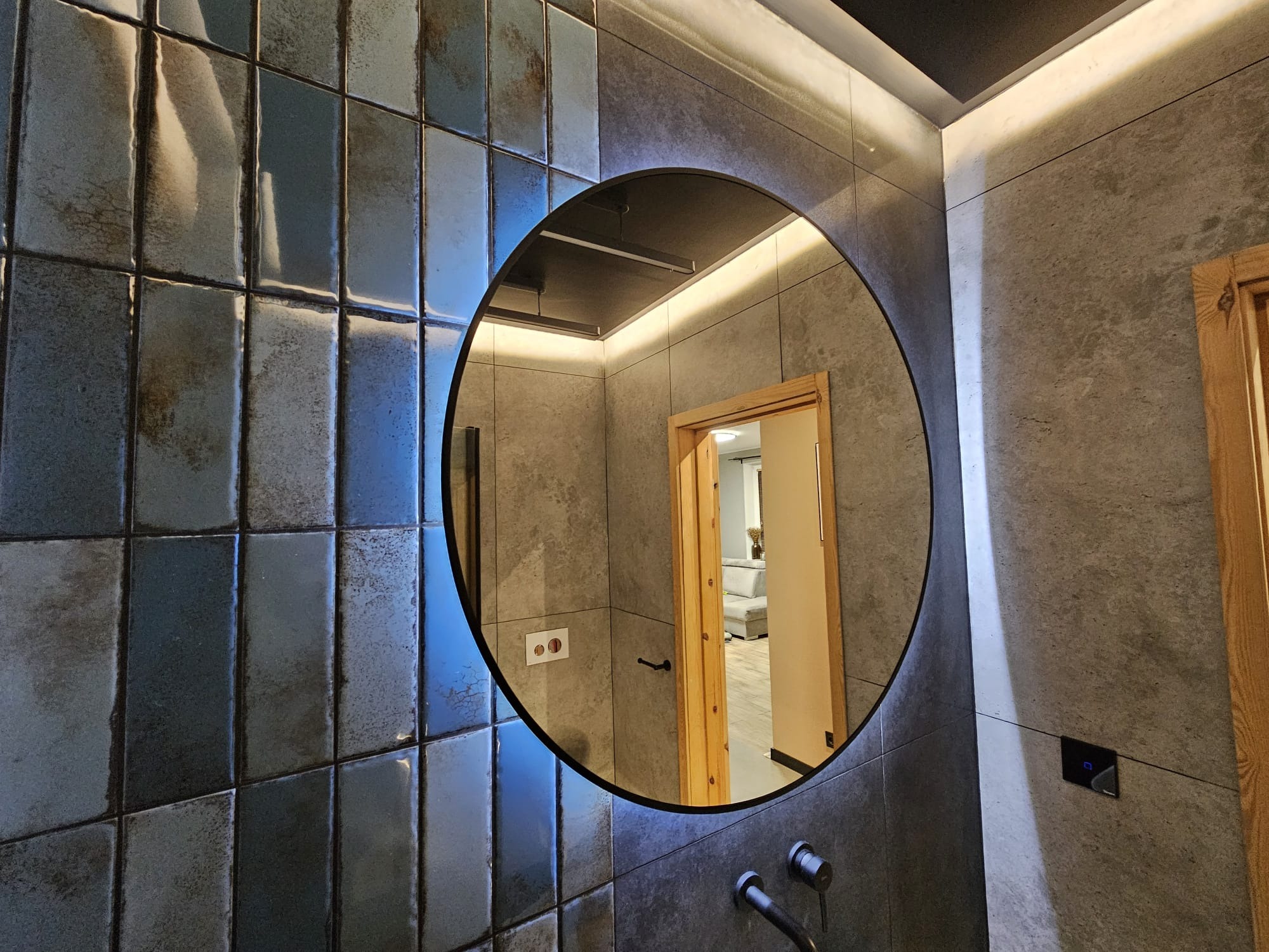 Łazienka z błyszczącymi, niebiesko-szarymi płytkami, okrągłym lustrem z podświetleniem LED i widocznym fragmentem wnętrza przez otwarte drzwi.