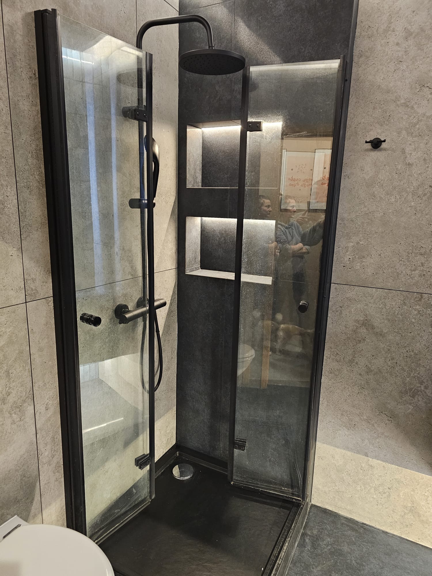 Nowoczesna kabina prysznicowa z czarnymi profilami, deszczownicą i wnękami z podświetleniem LED, widoczna w odbiciu sylwetka osoby i fragment toalety.