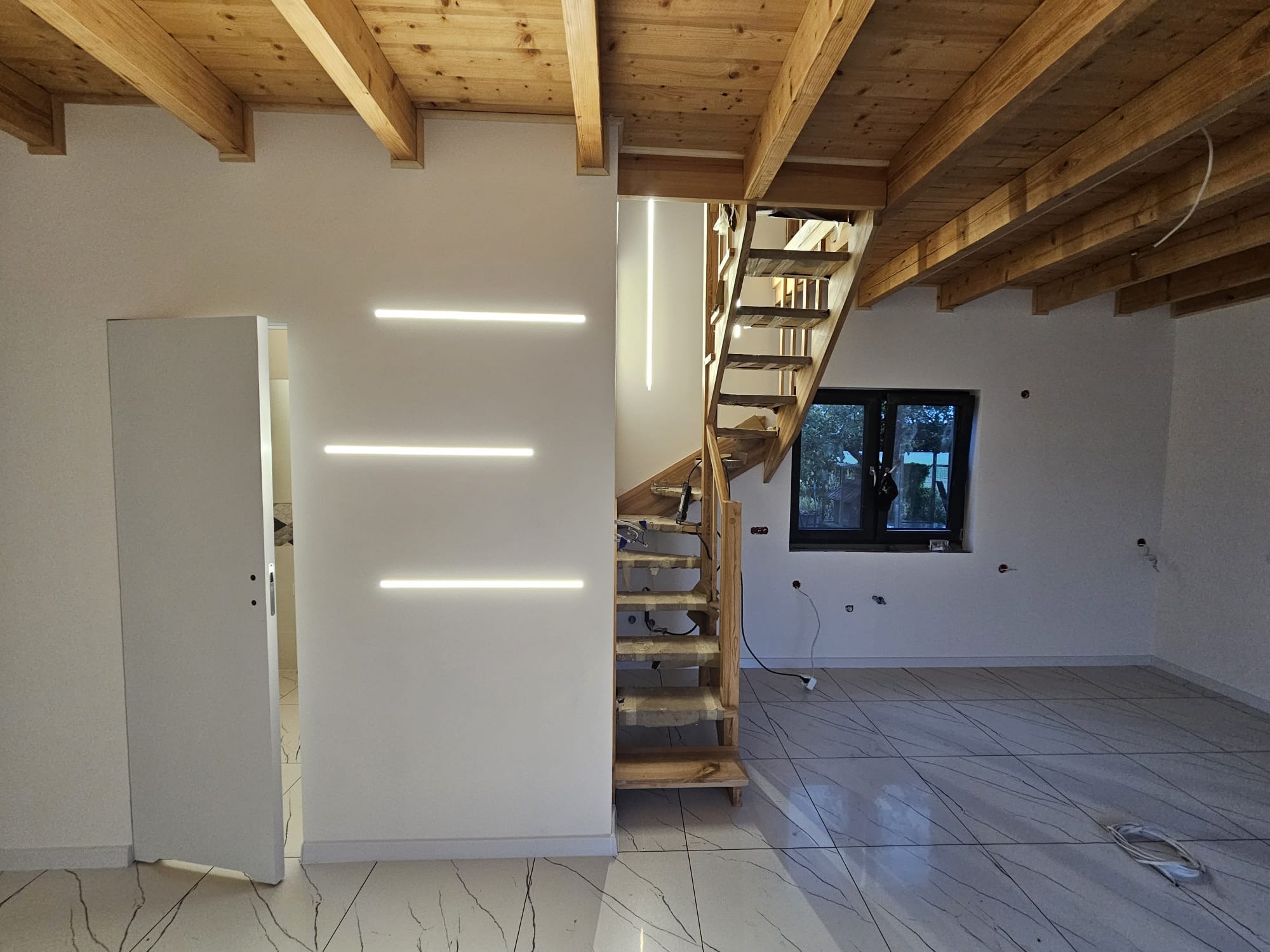 Wnętrze w trakcie prac wykończeniowych: drewniane schody bez balustrady prowadzą na górę, podświetlone poziomymi listwami LED na ścianie, widoczna drewniana konstrukcja sufitu, biała podłoga...