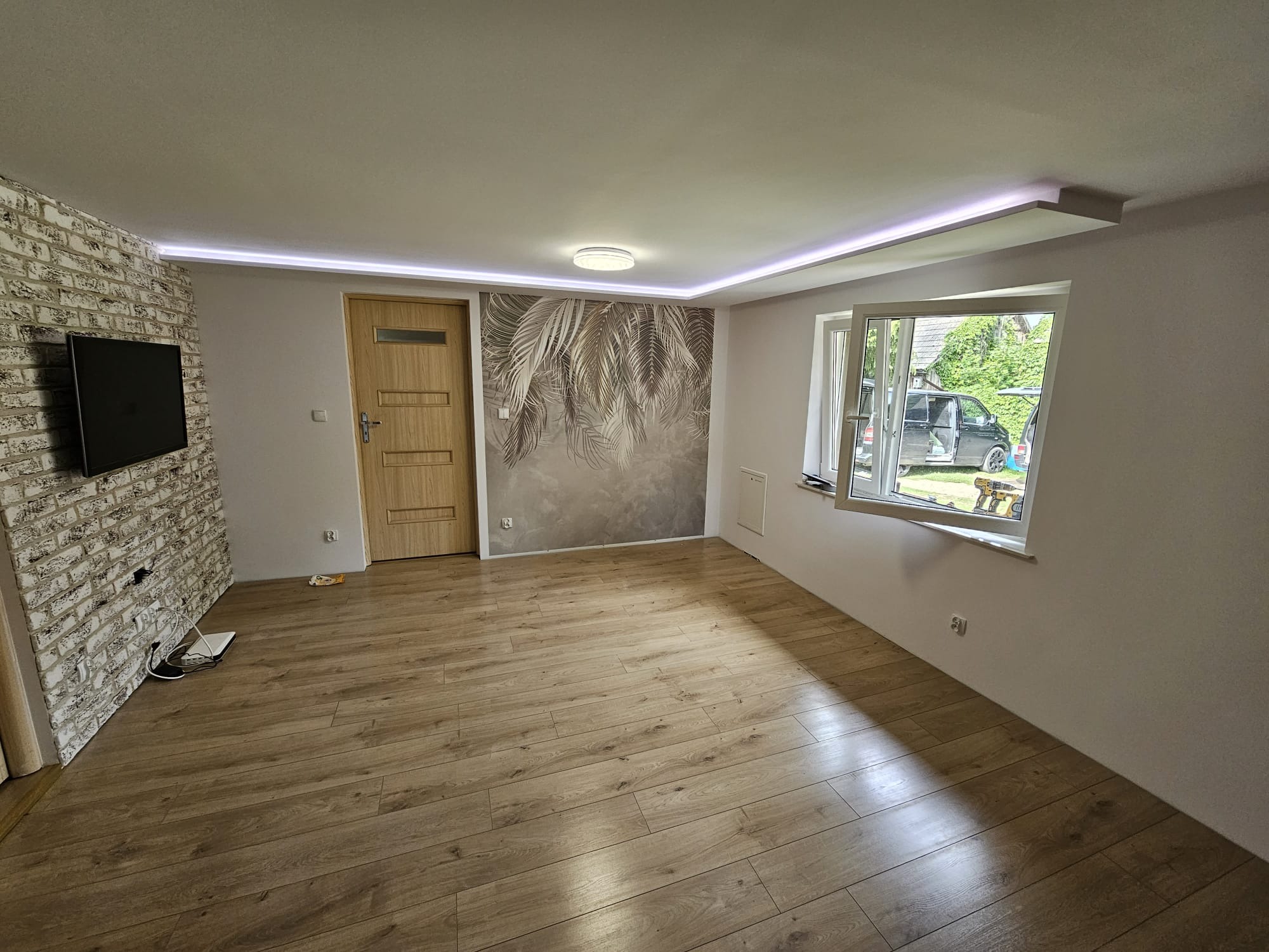 Pokój po renowacji z panelami podłogowymi w odcieniach drewna, ściana z cegły dekoracyjnej, tapeta z motywem roślinnym, oświetlenie LED podsufitowe oraz otwarte okno z widokiem na zewnątrz.